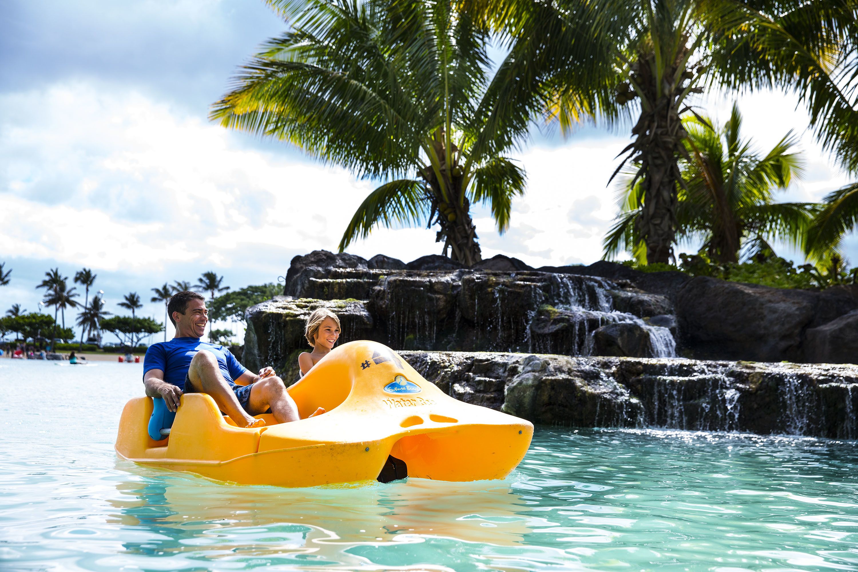 Ein Mann und eine Frau sitzen in einem gelben Luftboot in einem klaren, blauen Pool, im Hintergrund sind Palmen und ein Wasserfall zu sehen.
