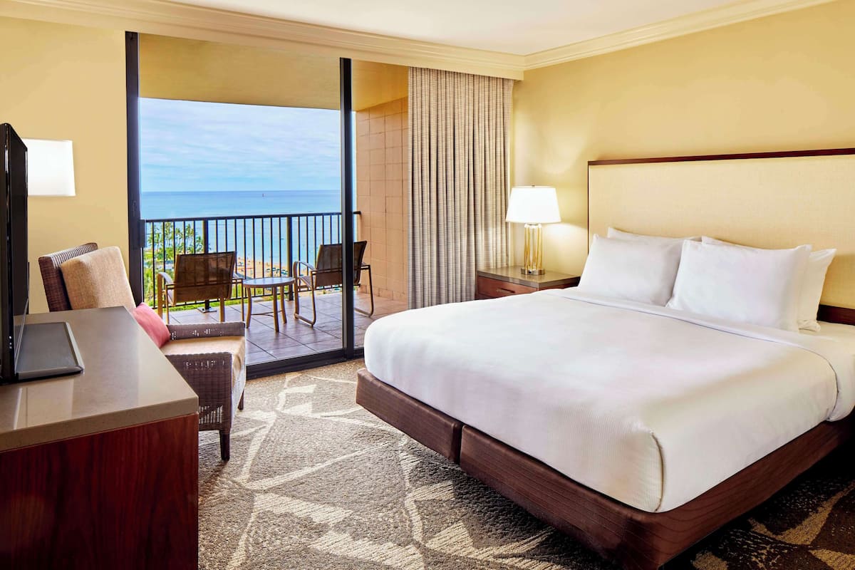 Habitación de hotel con una cama grande, un escritorio con una computadora portátil, una silla y vista al océano a través de una puerta corrediza.