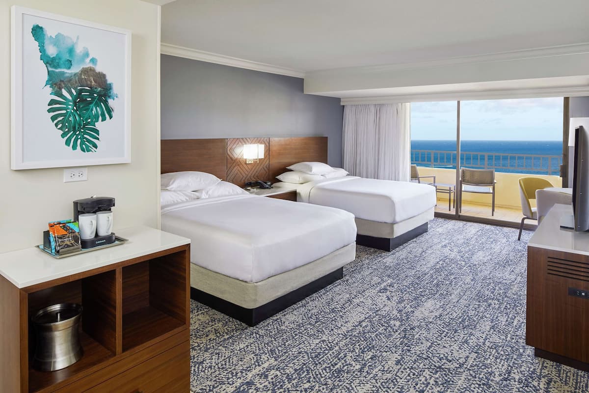 Habitación de hotel con dos camas, un escritorio, cafetera y vista al océano.