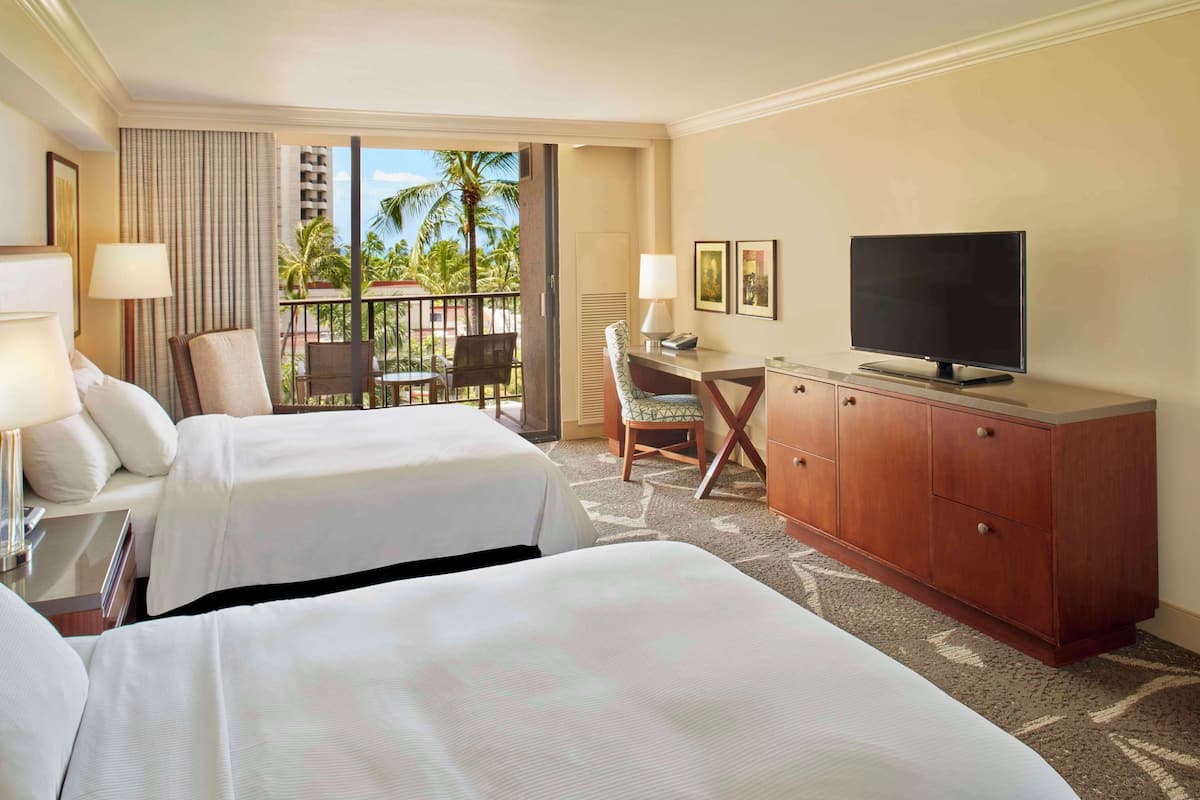 Habitación de hotel con dos camas, un escritorio con televisor, una silla y vistas a palmeras.