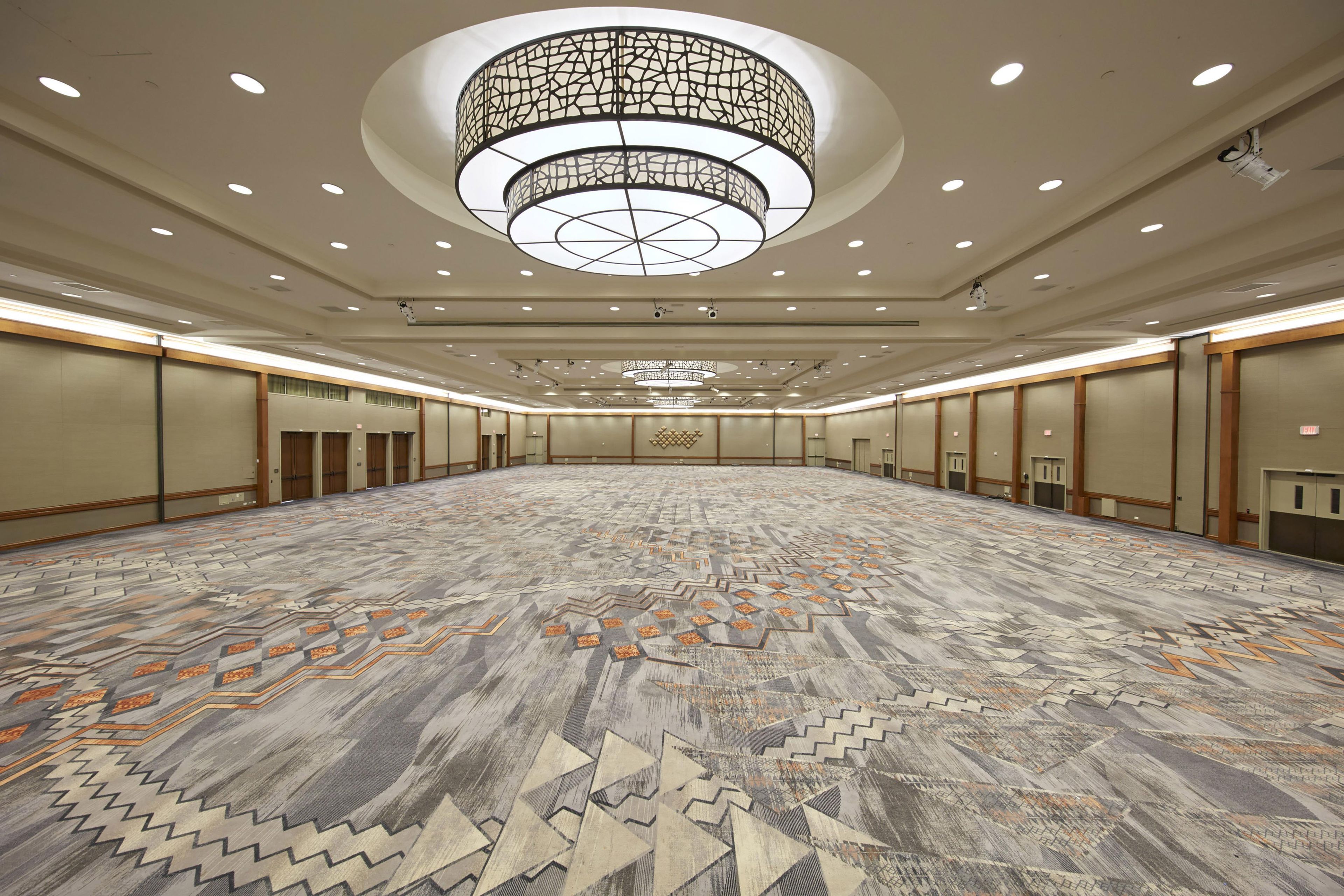 Une grande salle de conférence vide, dotée d’un tapis à motifs, d’un lustre central et de plusieurs portes le long des murs.