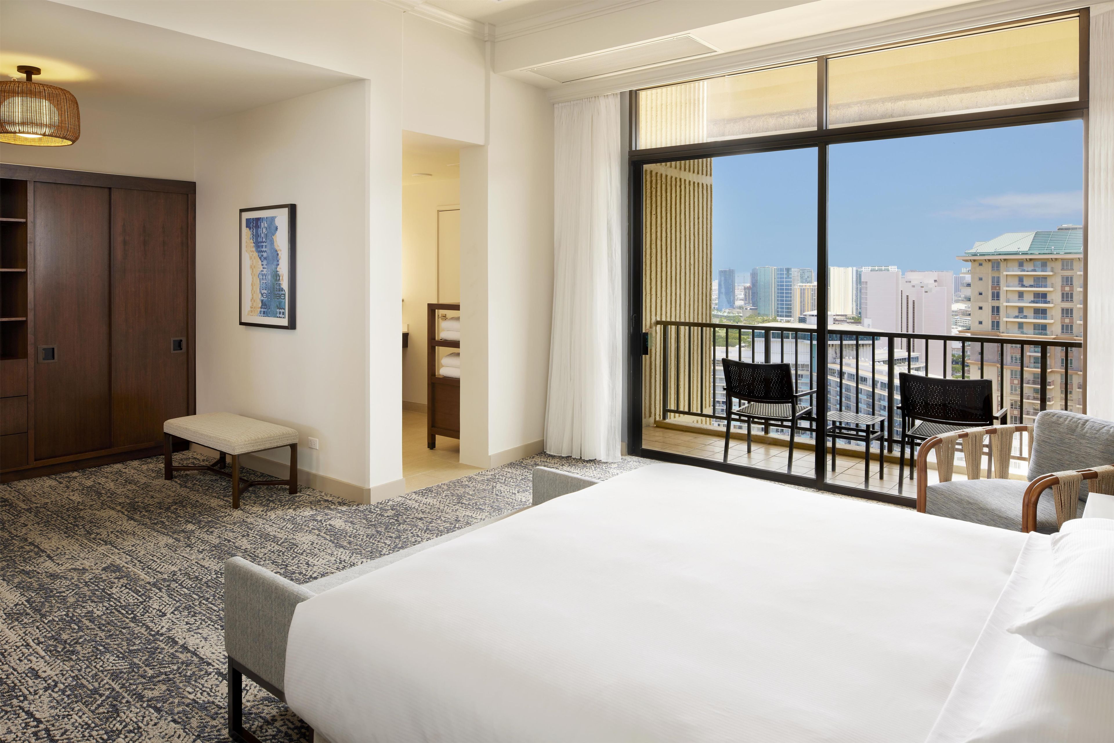 A hotel room with a large bed, a seating area with chairs, a view of the cityscape through a balcony, and a door leading to a bathroom.