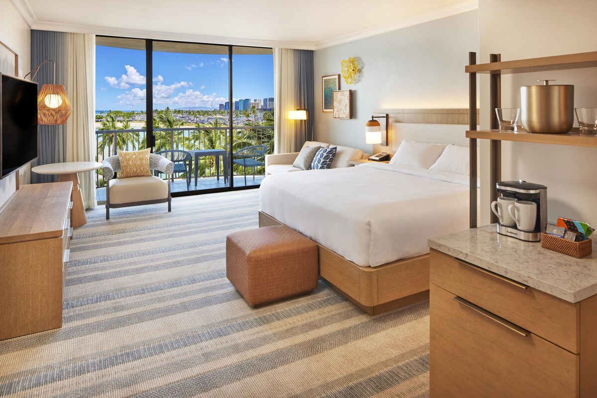 A hotel room with a large bed, a TV, a balcony with a city view, and a breakfast bar.
