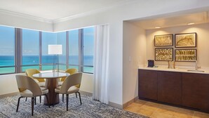 A hotel room with a round dining table, four chairs, a large window with a view of the ocean, a kitchen area with a counter and cabinets, and a patterned rug.