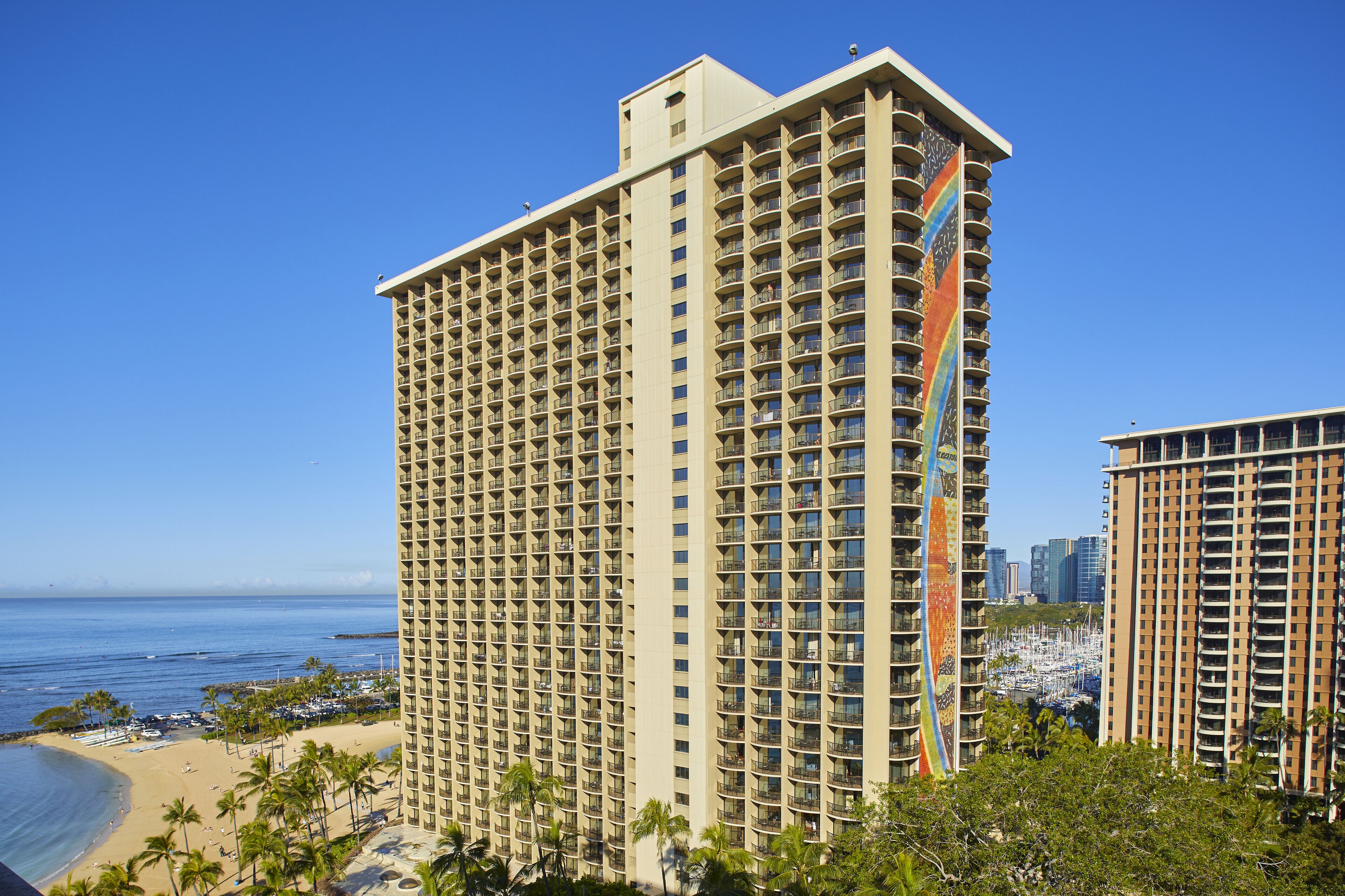 A tall hotel with multiple balconies, located near a beach with palm trees and a marina.
