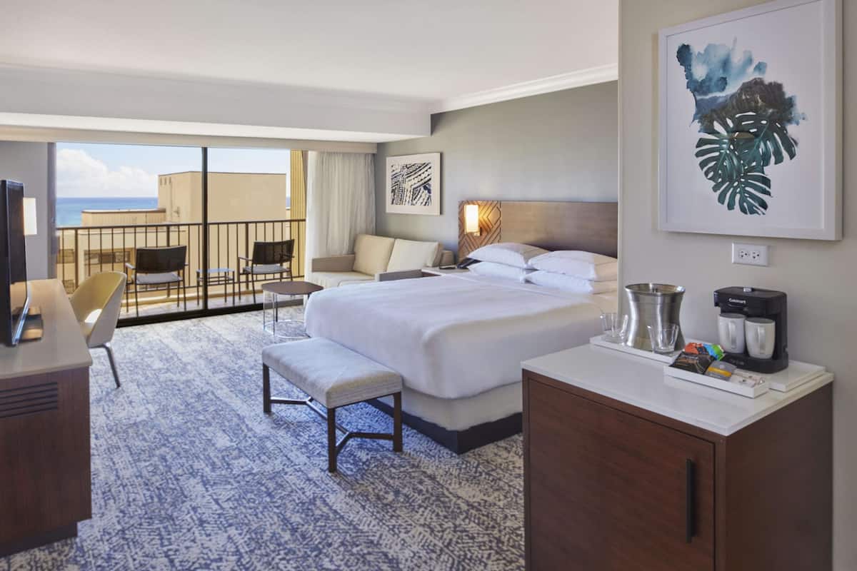 Habitación de hotel con una cama grande, un escritorio con computadora, una silla, una mesita y vista al océano.