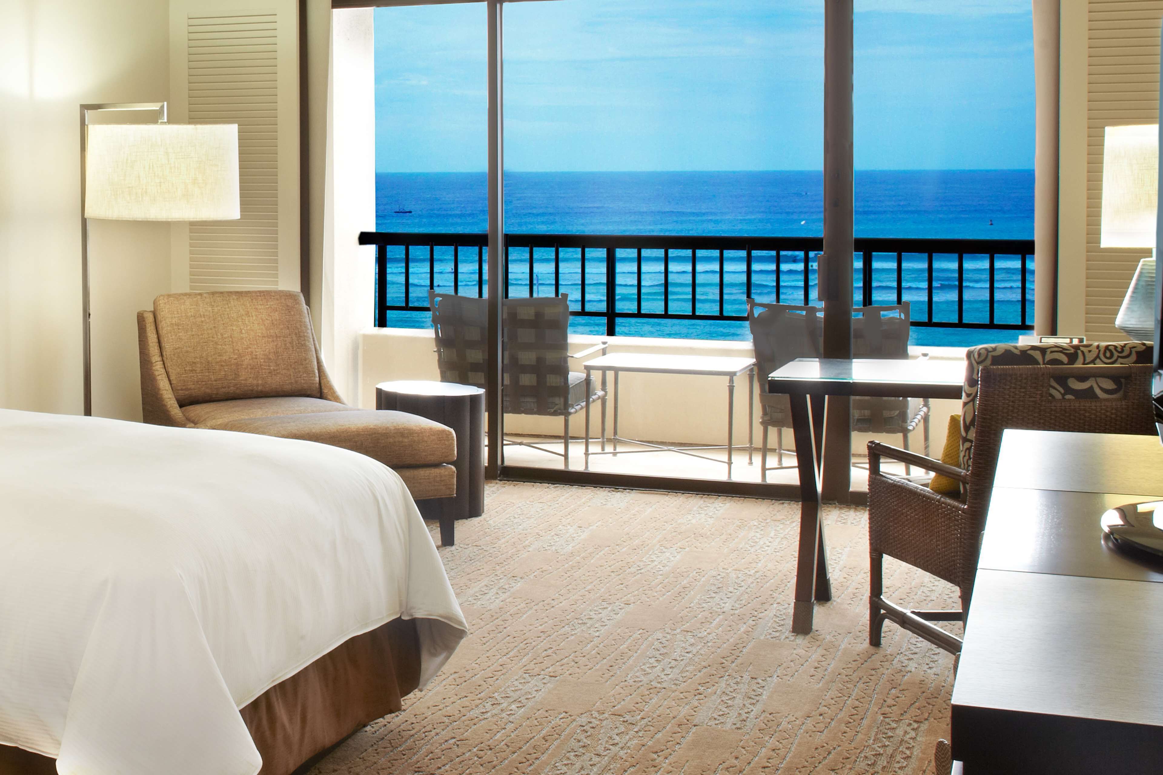 A hotel room with a large bed, a sitting area with a table and chairs, and a balcony with a view of the ocean.