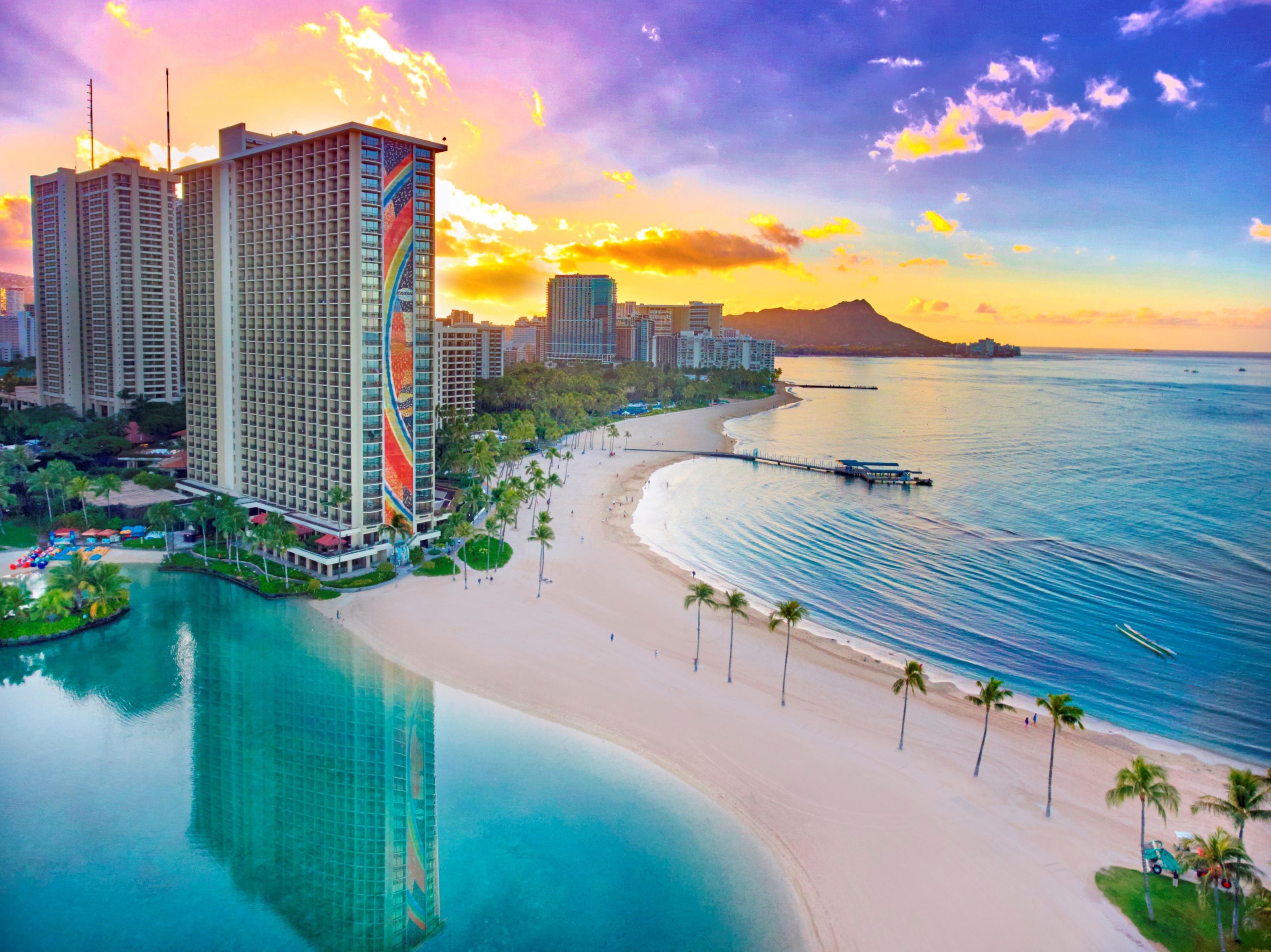 A coastal hotel with a beach view, a pool, and a city skyline in the background.