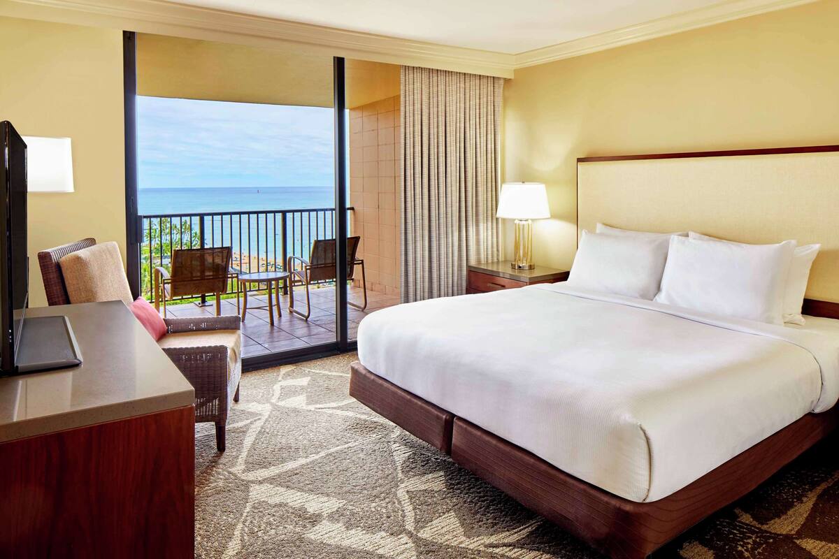 Habitación de hotel con una cama grande, un escritorio con una computadora portátil, una silla y vista al océano a través de una puerta corrediza.