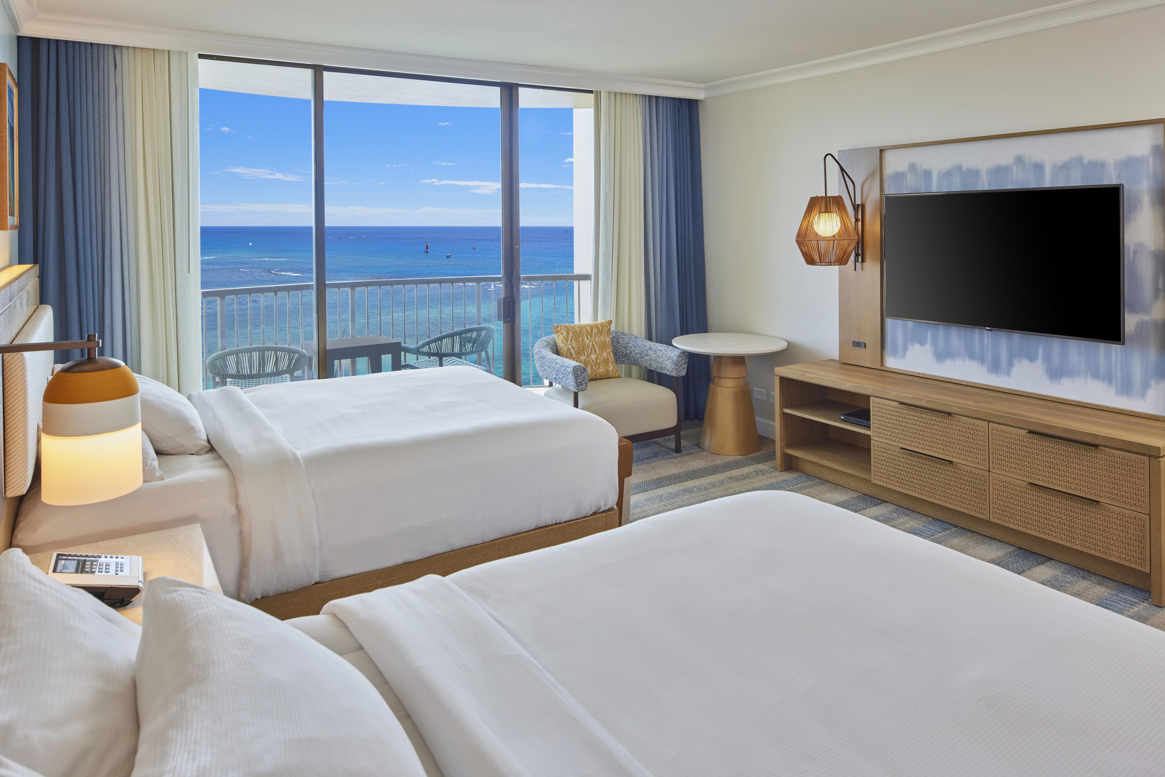 Een hotelkamer met twee bedden, een televisie, een kleintje tafeltje en een balkon met uitzicht op de oceaan.