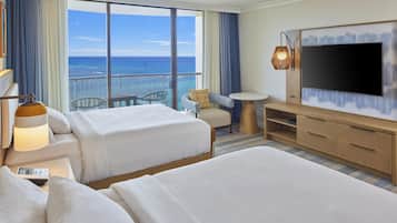 Habitación de hotel con dos camas, televisión, una mesita y balcón con vista al océano.