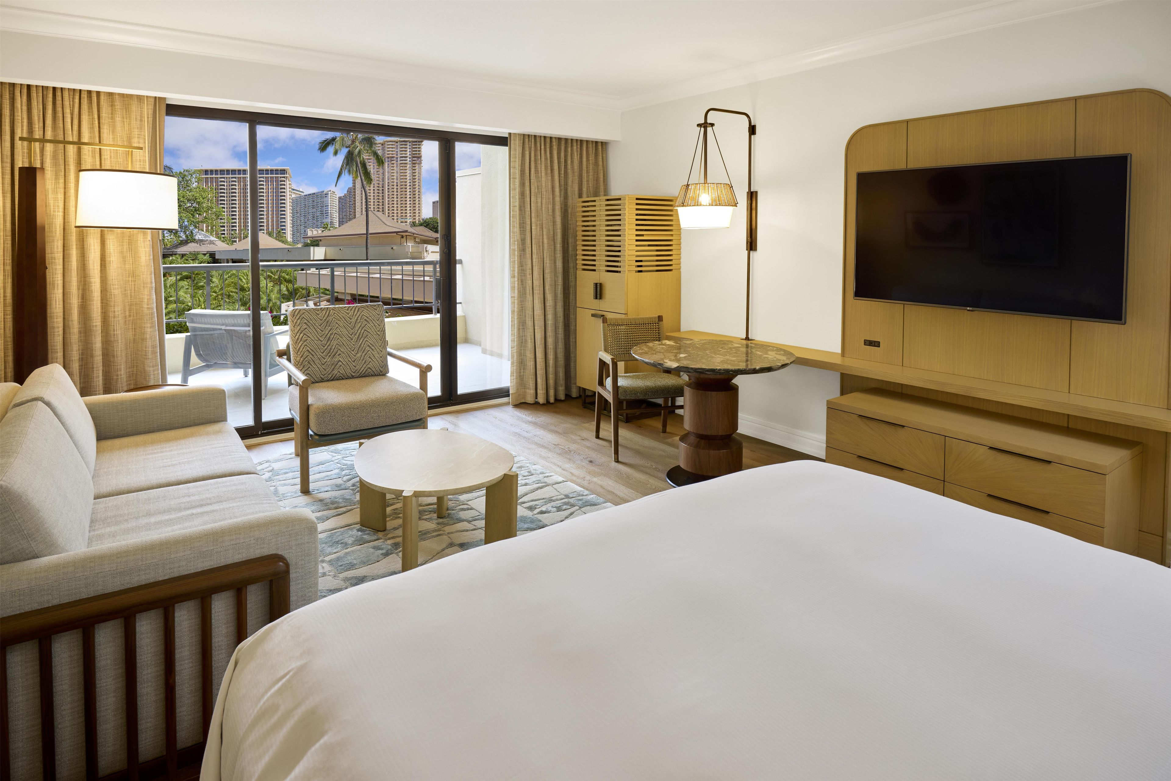 Habitación de hotel con cama, sofá, mesa de comedor, silla, televisión y balcón con vistas a edificios.