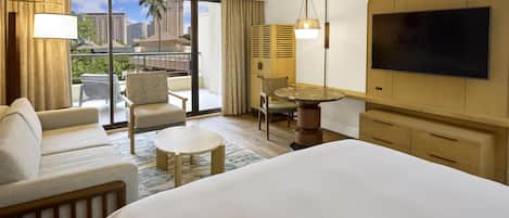 Habitación de hotel con cama, sofá, mesa de comedor, silla, televisión y balcón con vistas a edificios.