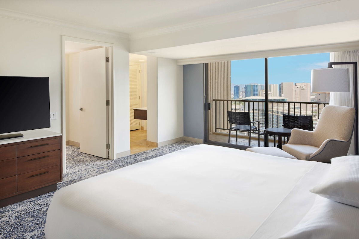 Habitación de hotel con una cama grande, televisión, escritorio y balcón con vistas a la ciudad.