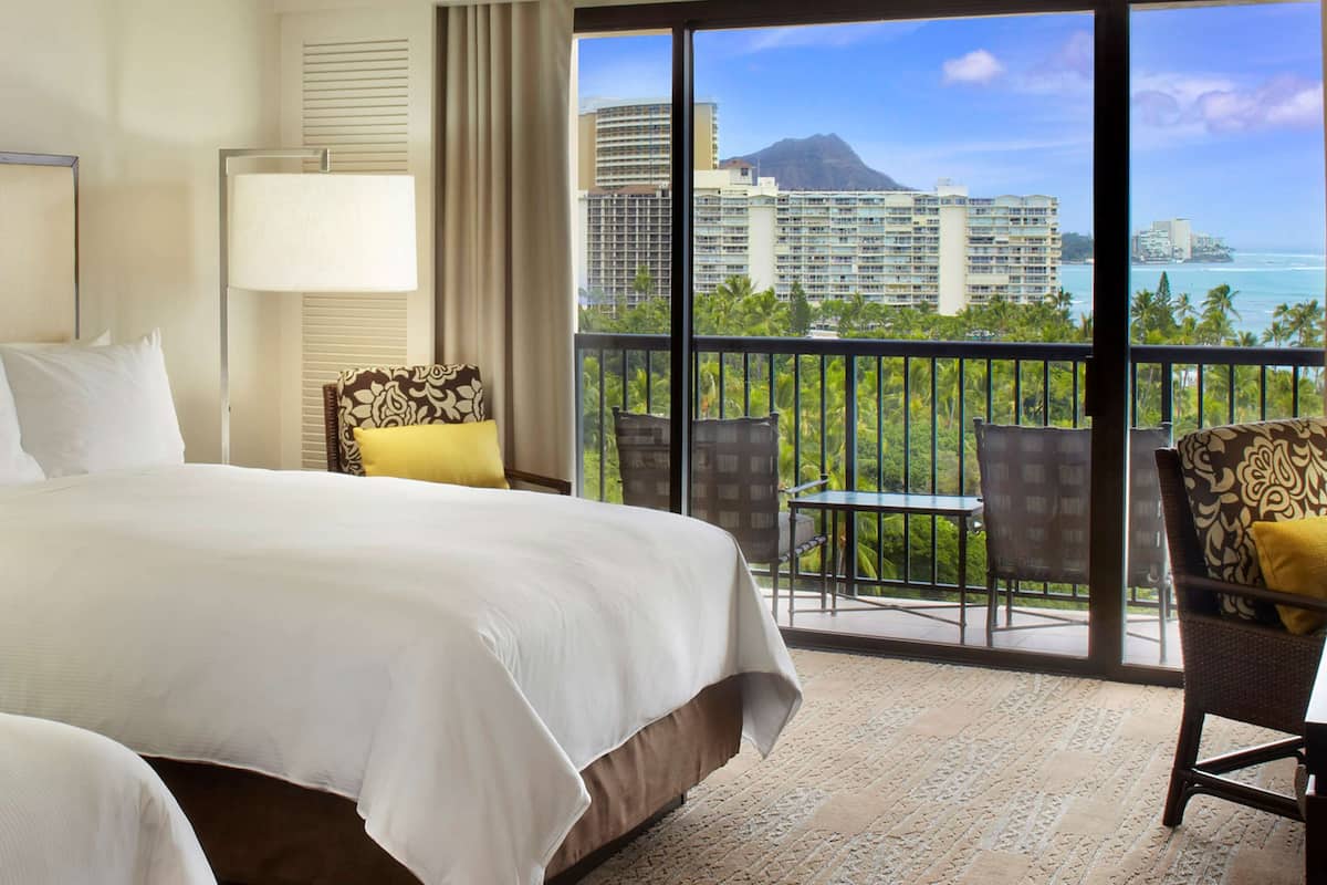 Habitación de hotel con una cama grande, un balcón con mesa y sillas, y vistas a la ciudad y al océano.