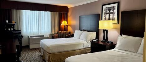 A hotel room with two beds, a desk, a chair, a lamp, an air conditioner, and a framed picture on the wall.