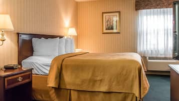 A hotel room with a large bed, two bedside lamps, a nightstand with a radio, a framed picture on the wall, and a window with sheer curtains.