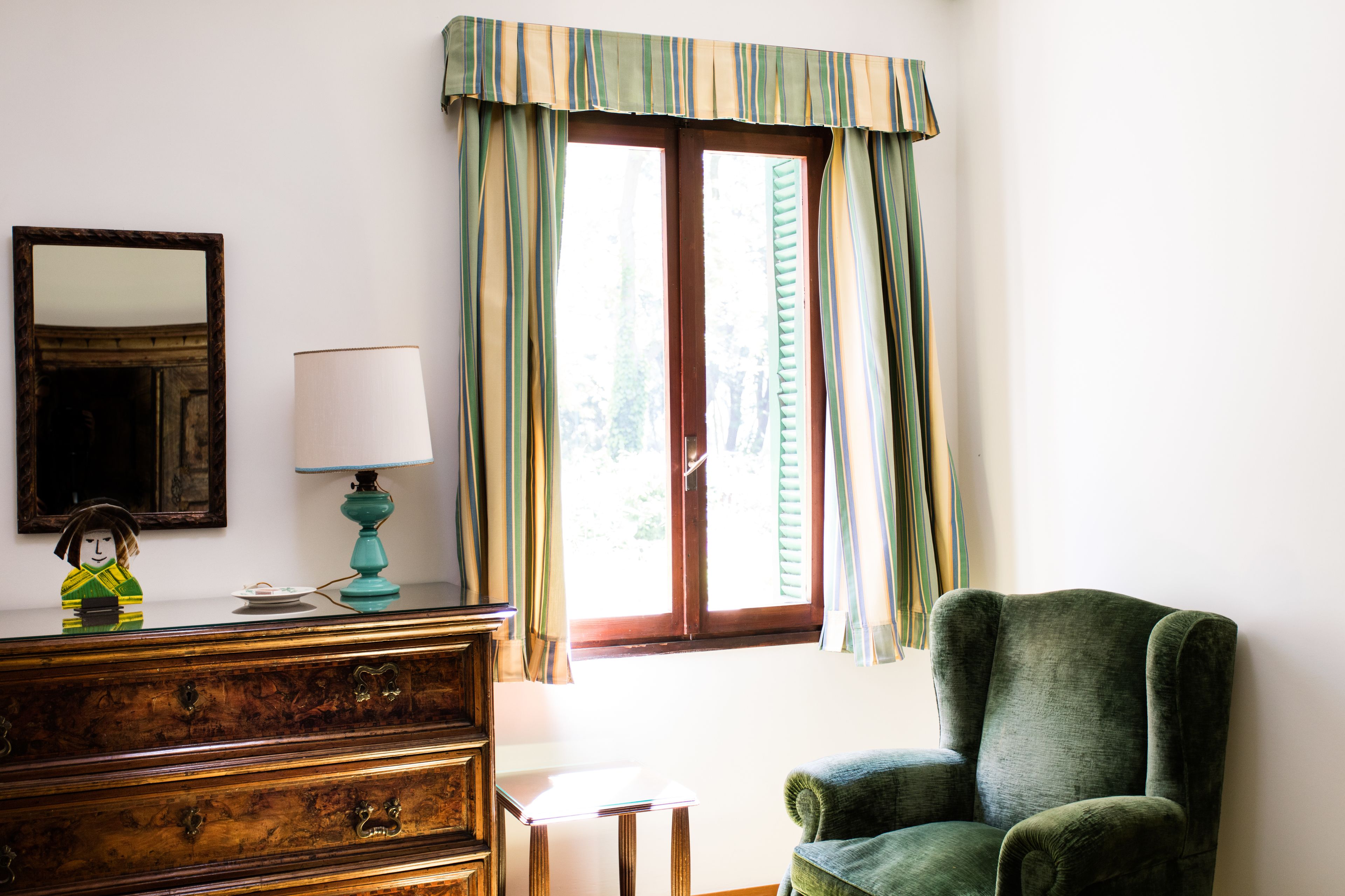 A room with a wooden dresser, a lamp, a chair, a window with curtains, and a mirror on the wall.