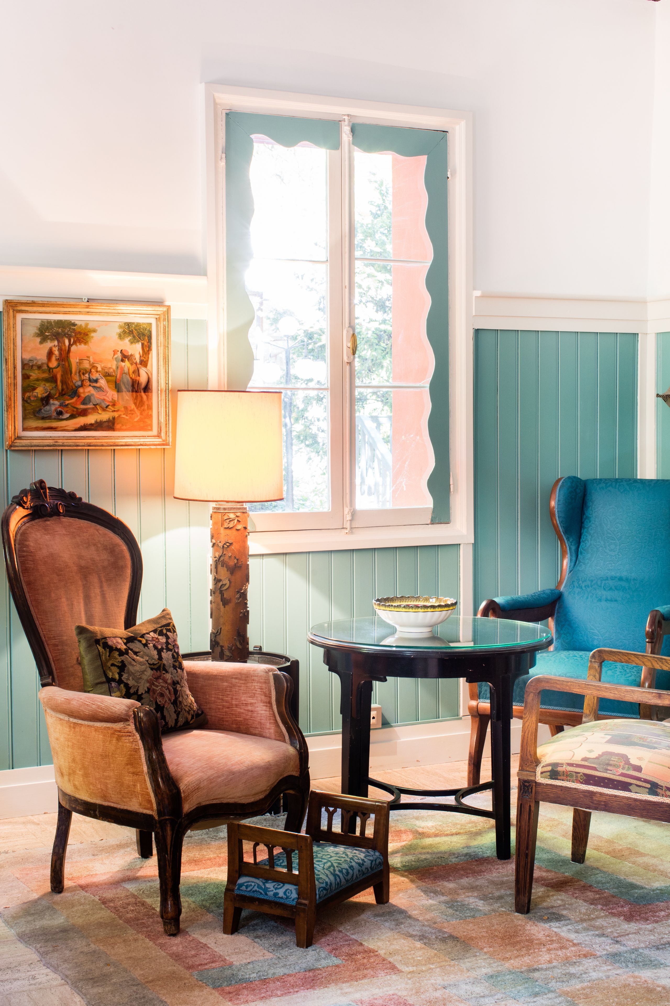 A room with a teal wall, a window with a wavy design, a lamp, a chair, a table, and a small rug.