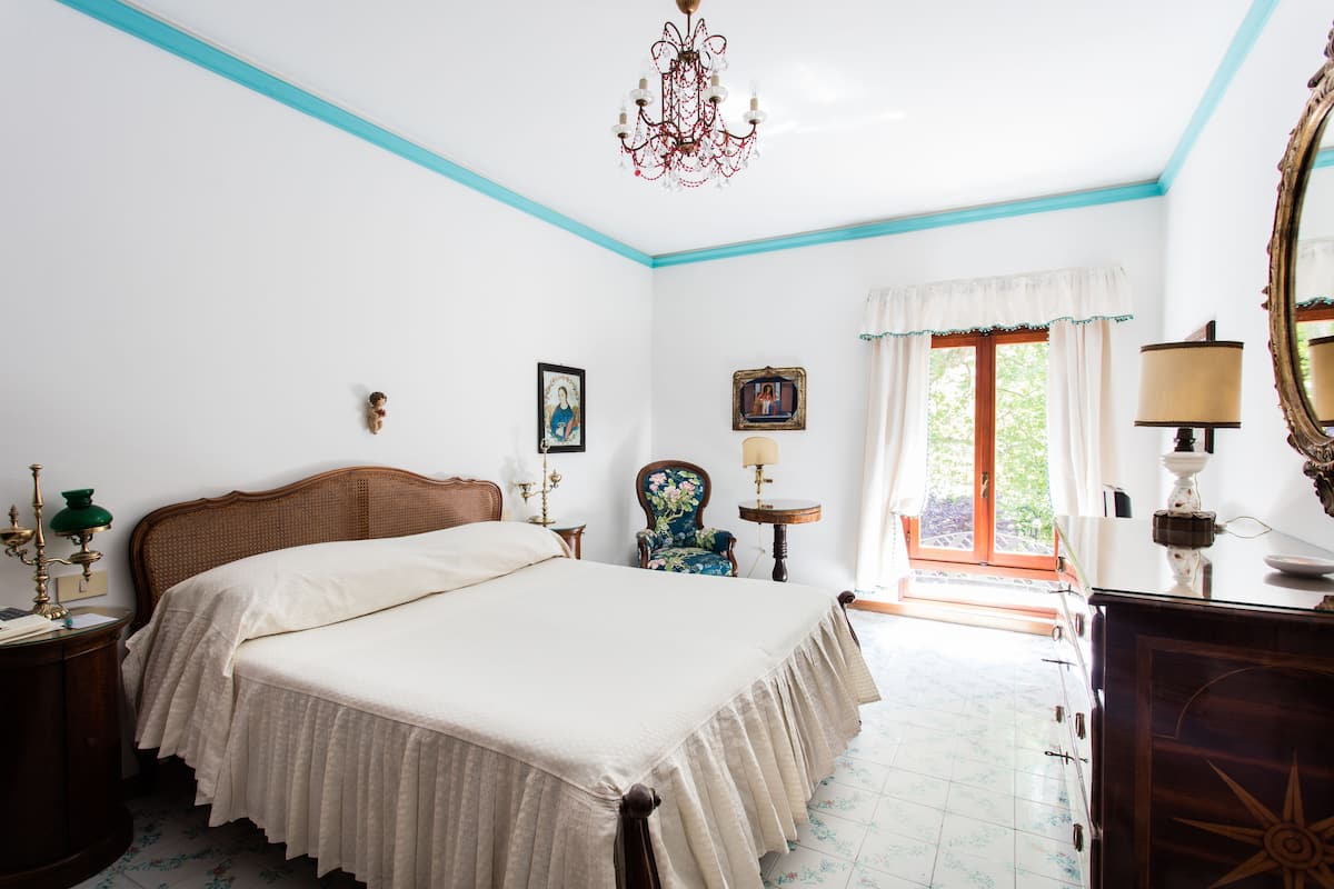 A bedroom with a large bed, a wooden headboard, a chandelier, a chair, a table with a lamp, and a window with curtains.
