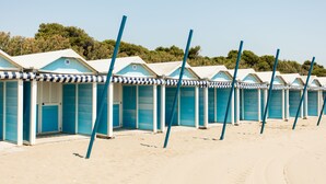 Una hilera de cabañas de playa con puertas azules y toldos a rayas, alineadas en una playa arenosa.