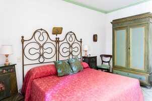 Quarto com estrutura de cama em metal, colcha vermelha, duas almofadas decorativas, um armário verde e uma cadeira de madeira.