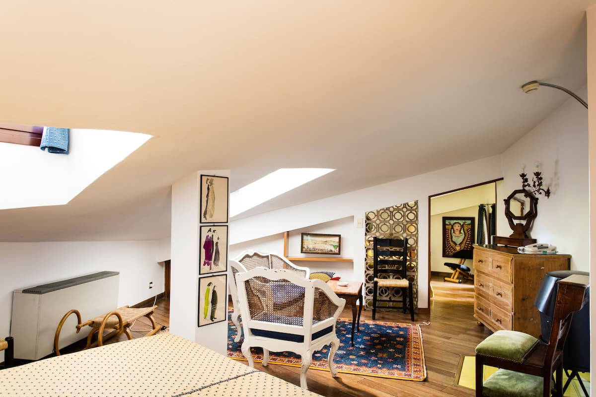 A room with a sloped ceiling, a wooden floor, a rocking chair, a dining table, a dresser, and a large window.