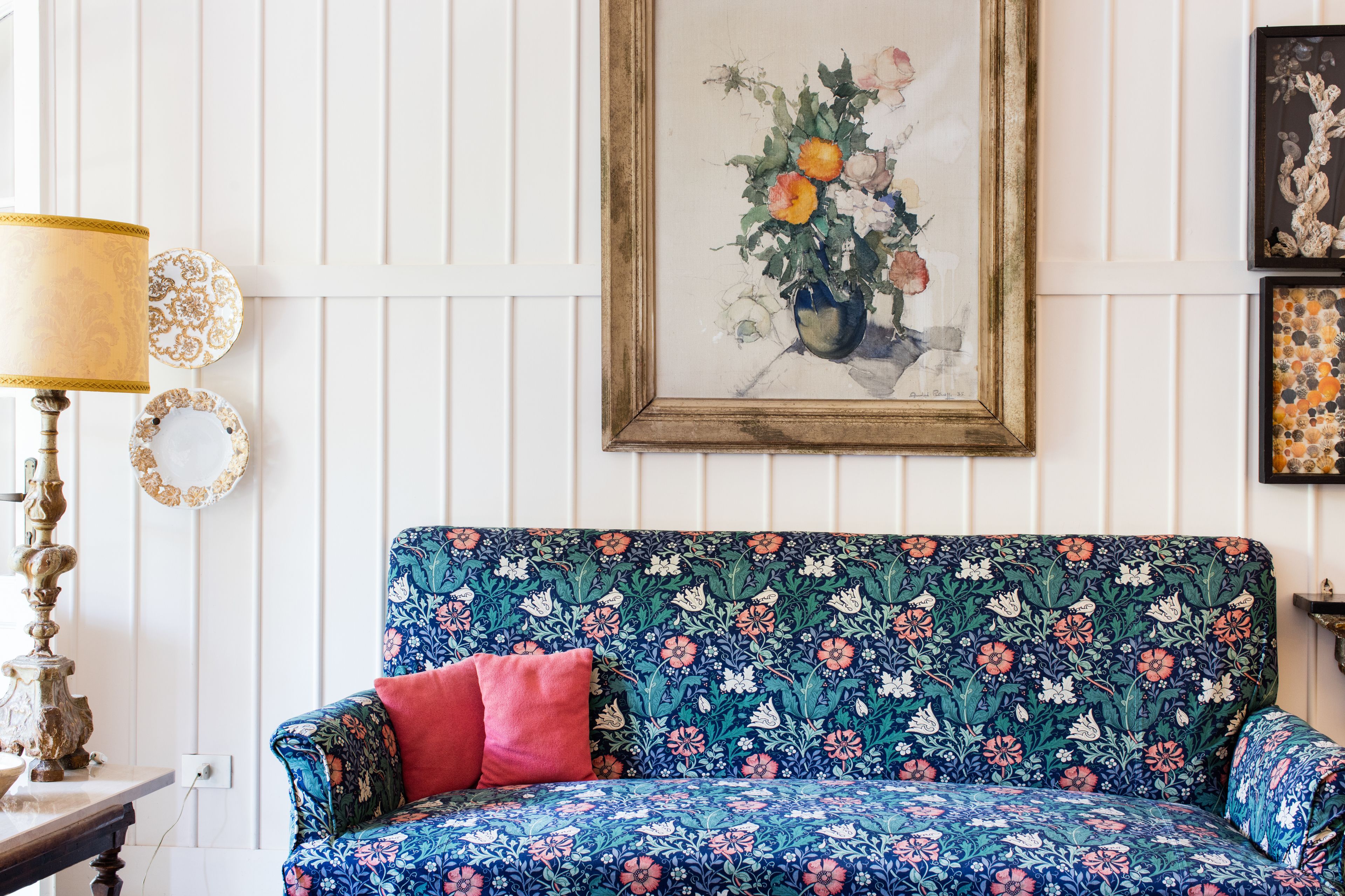 Un sofá estampado con motivos florales, un cojín rojo y una lámpara antigua.