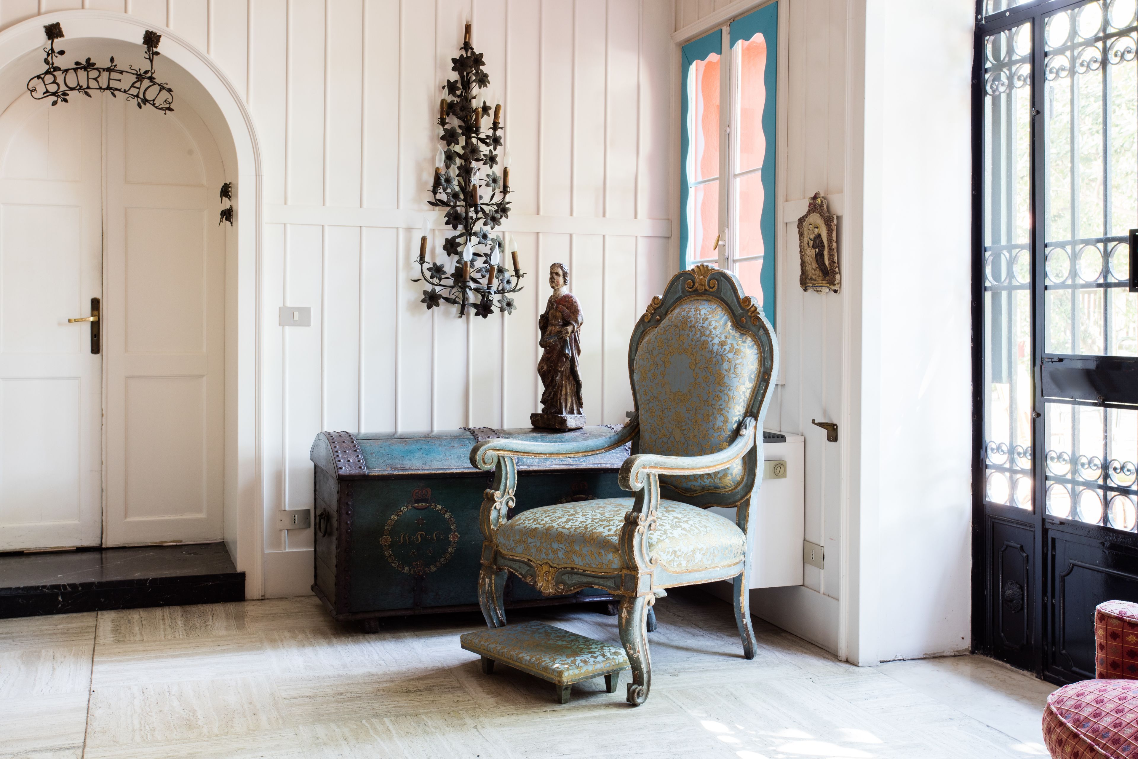 A room with a white door, a blue armchair, a small table, and a decorative wall piece.