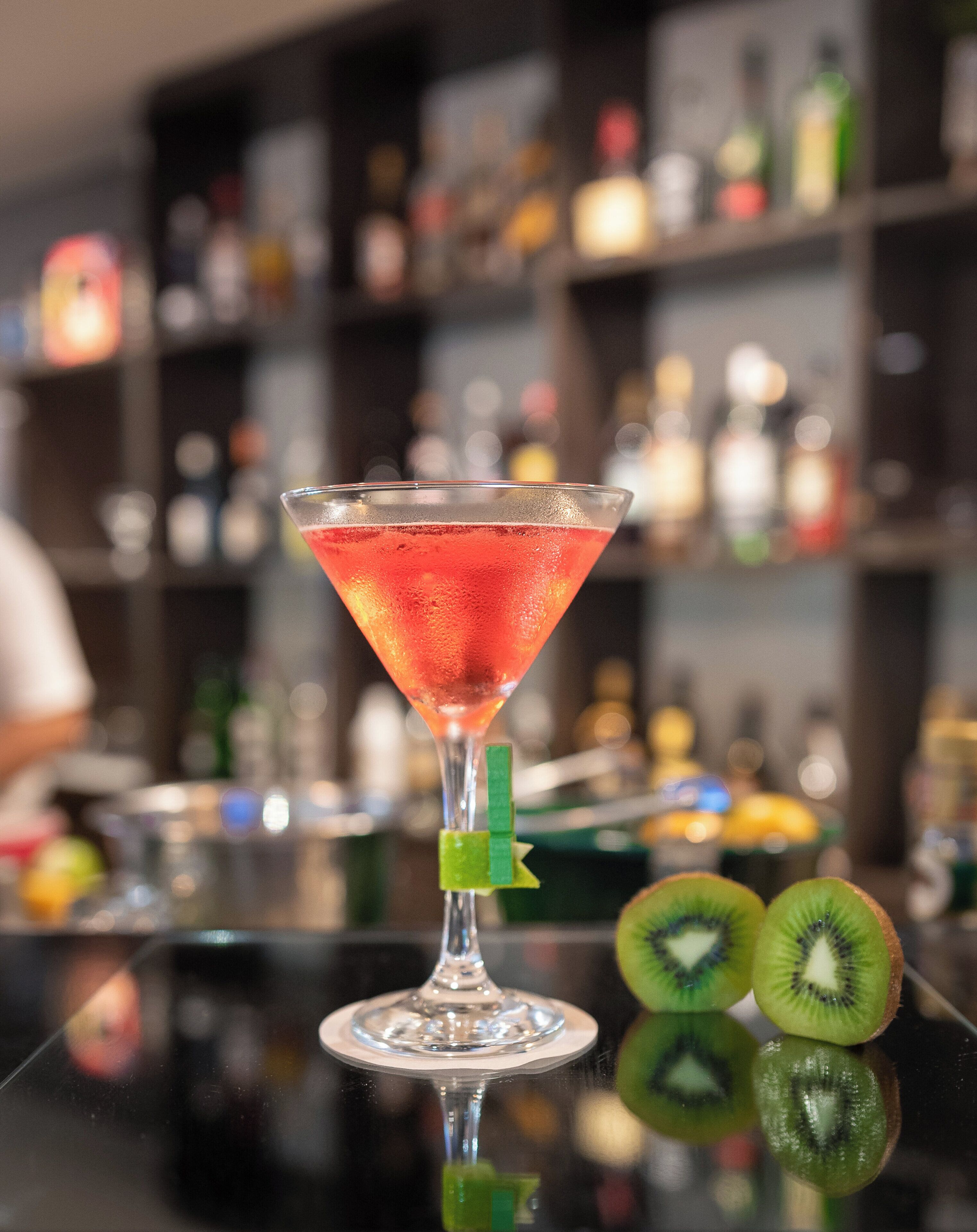 A martini glass with a red cocktail on a bar counter, accompanied by kiwi slices.