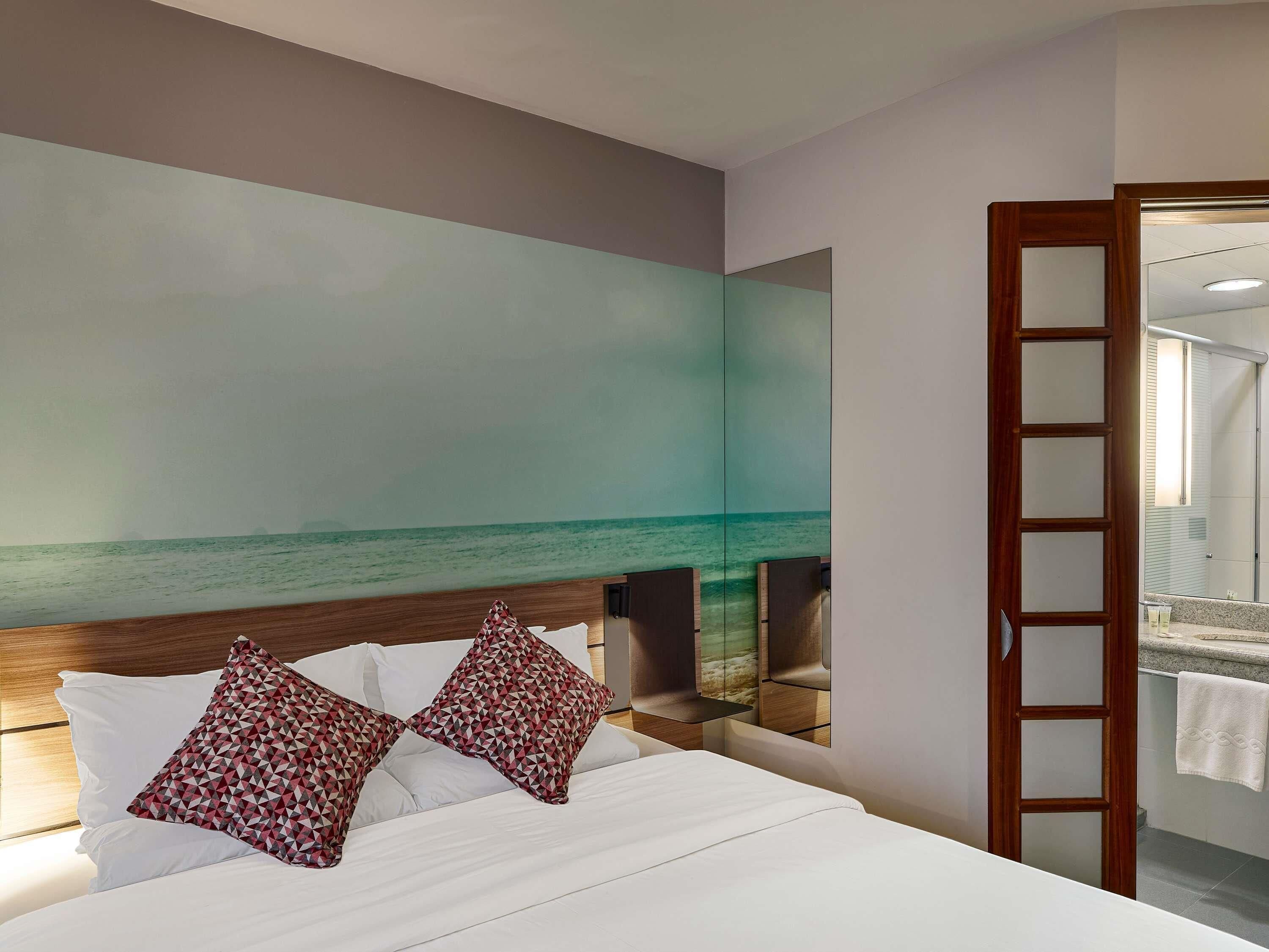 A hotel room with a bed, a large wall mural of a beach, a wooden headboard, and a mirror.
