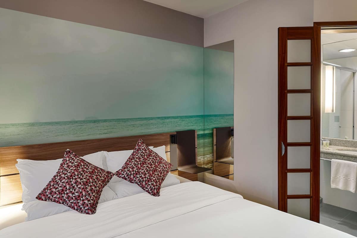A hotel room with a bed, a large wall mural of a beach, a wooden headboard, and a mirror.