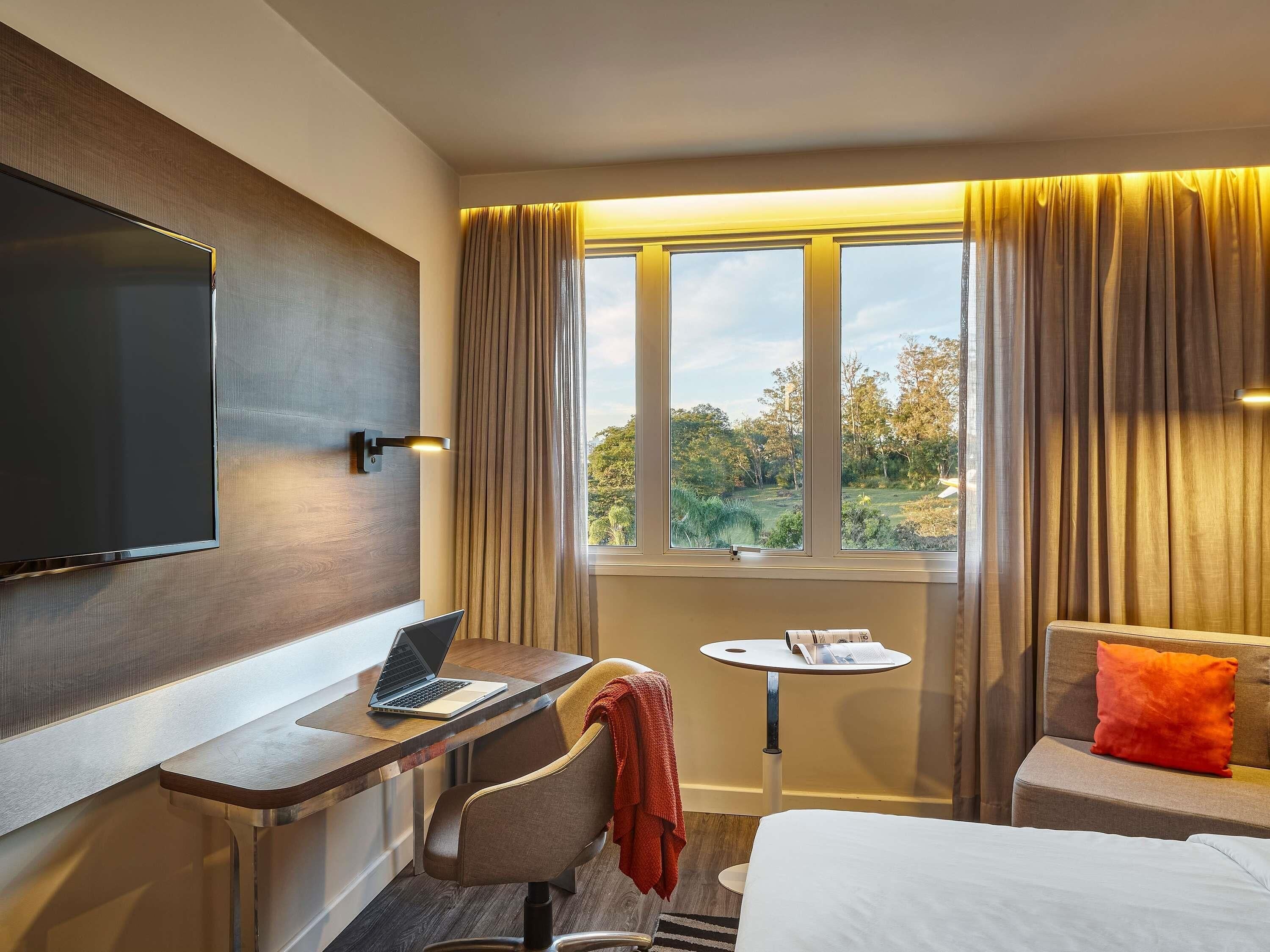 A hotel room with a bed, a desk with a laptop, a small table with a book, and a TV mounted on the wall.