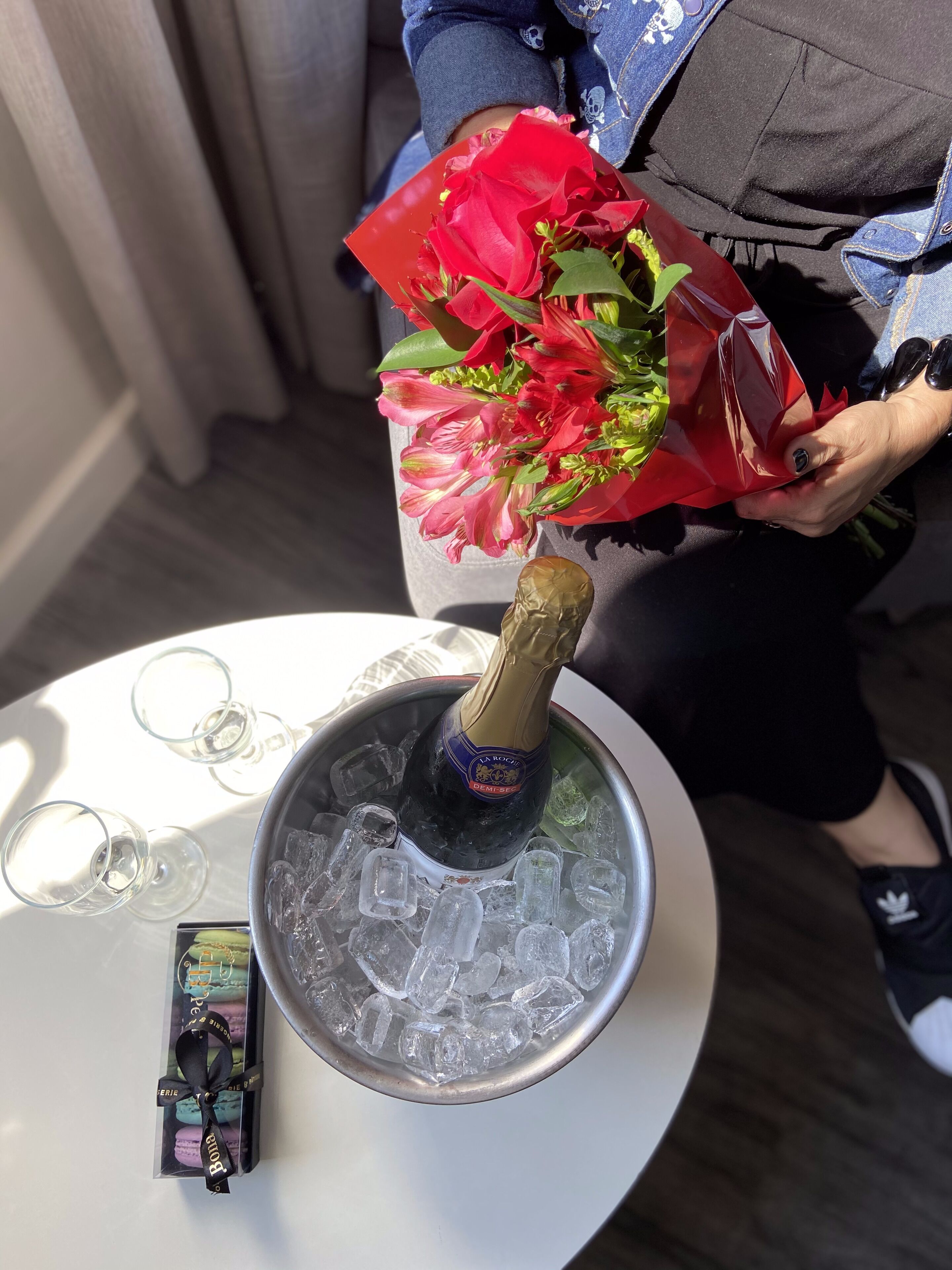 Una botella de champán en un balde con cubitos de hielo, un ramo de flores rojas y una caja de macarons sobre una mesa.