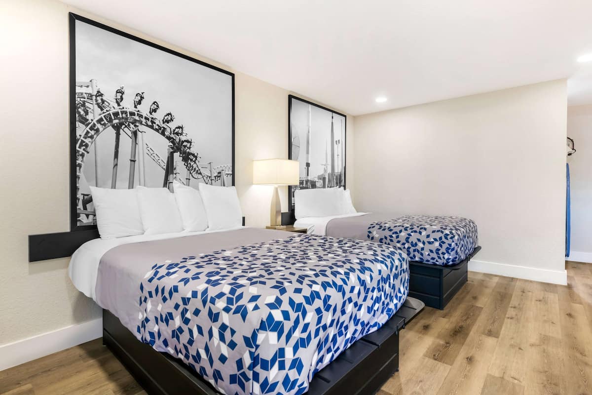 Habitación de hotel con dos camas, un cuadro grande enmarcados de una montaña rusa, una lámpara y vistas a un edificio.