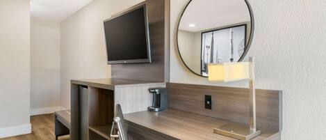 A modern hotel room with a wooden desk, a flat-screen TV, a round mirror, and a bedside lamp.