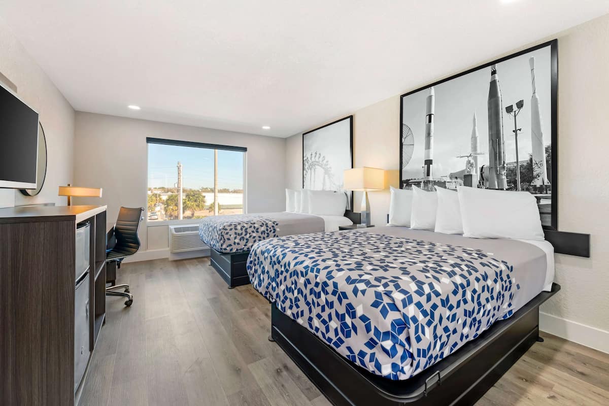 Habitación de hotel con dos camas, un escritorio, una silla, un televisor y un ventanal grande.