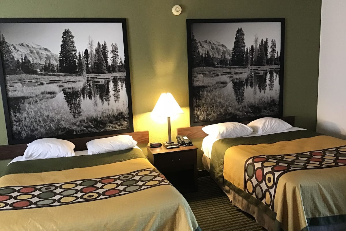 A hotel room with two beds, a nightstand with a lamp, and two framed landscape paintings on the walls.