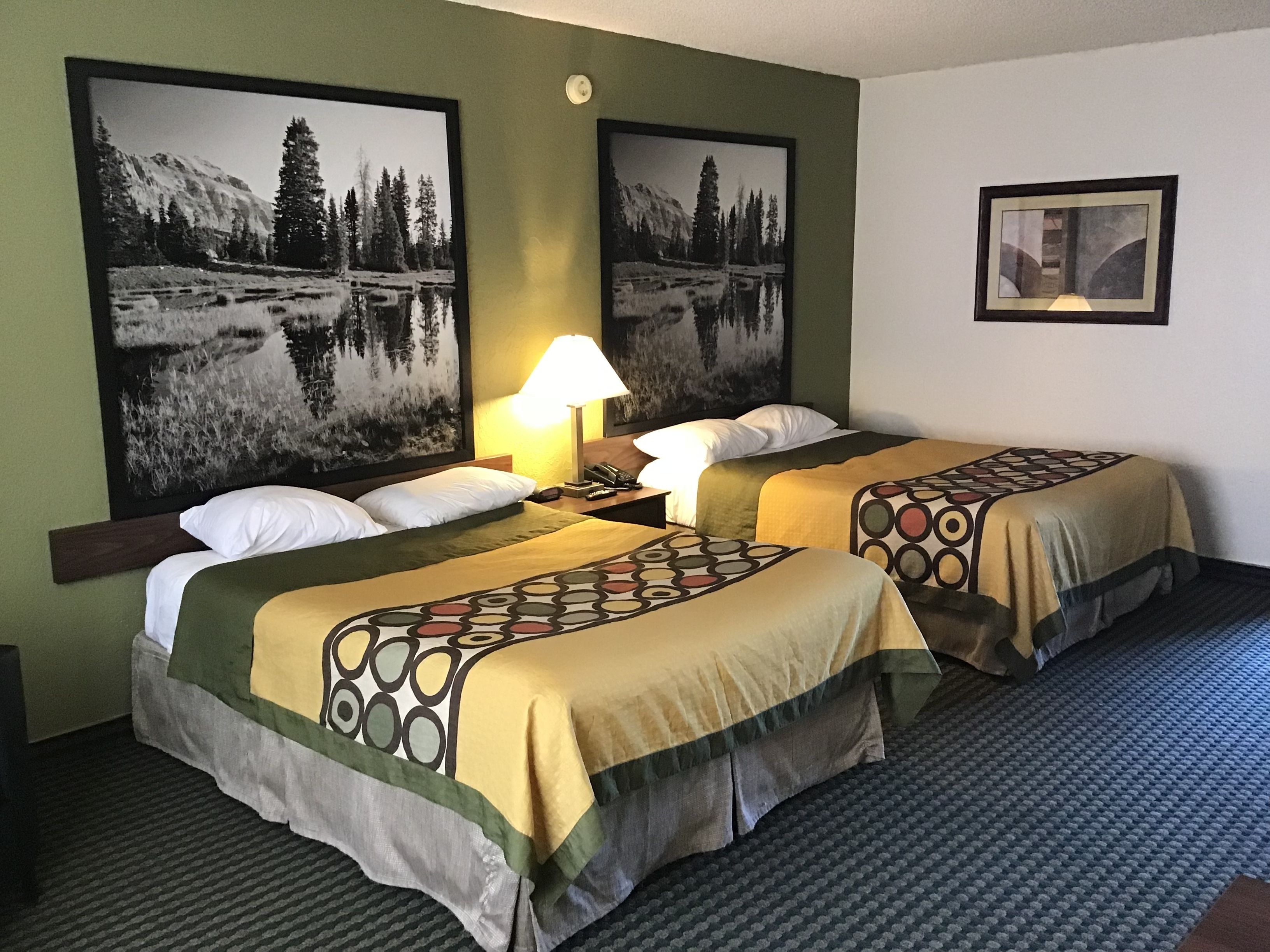 A hotel room with two beds, each with a patterned bedspread, a nightstand with a lamp, and two large framed pictures of a mountainous landscape.