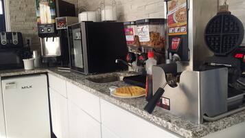 A kitchen with a milk fridge, coffee machine, waffle maker, and a waffle on a plate.