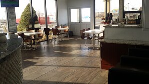 Um café espaçoso com piso de madeira, várias mesas e cadeiras, uma grande janela com vista para a rua, e um balcão de café com diversas bebidas e doces.