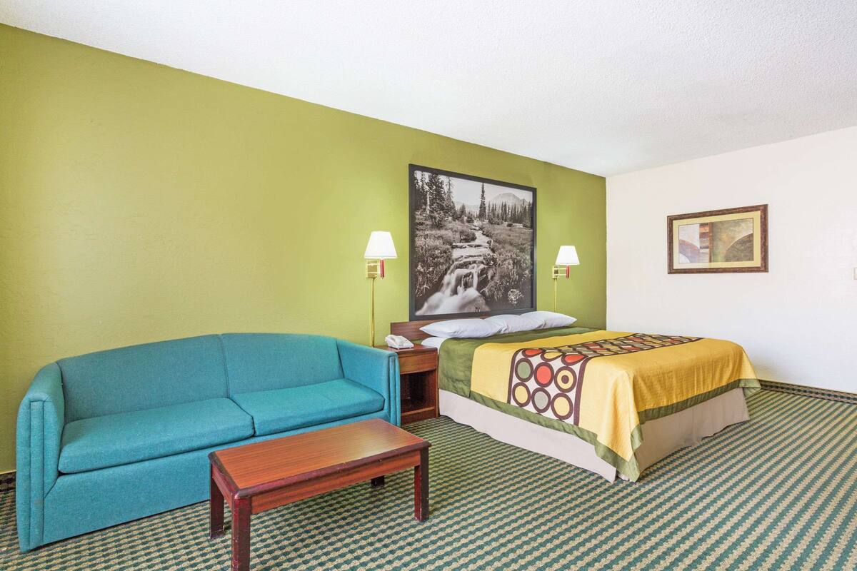 A hotel room with a green wall, a blue sofa, a wooden table, a bed with a patterned bedspread, and two wall-mounted lamps.