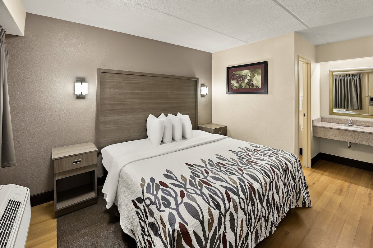 A hotel room with a bed, bedside tables, a nightstand, a mirror, and a bathroom with a sink and mirror.