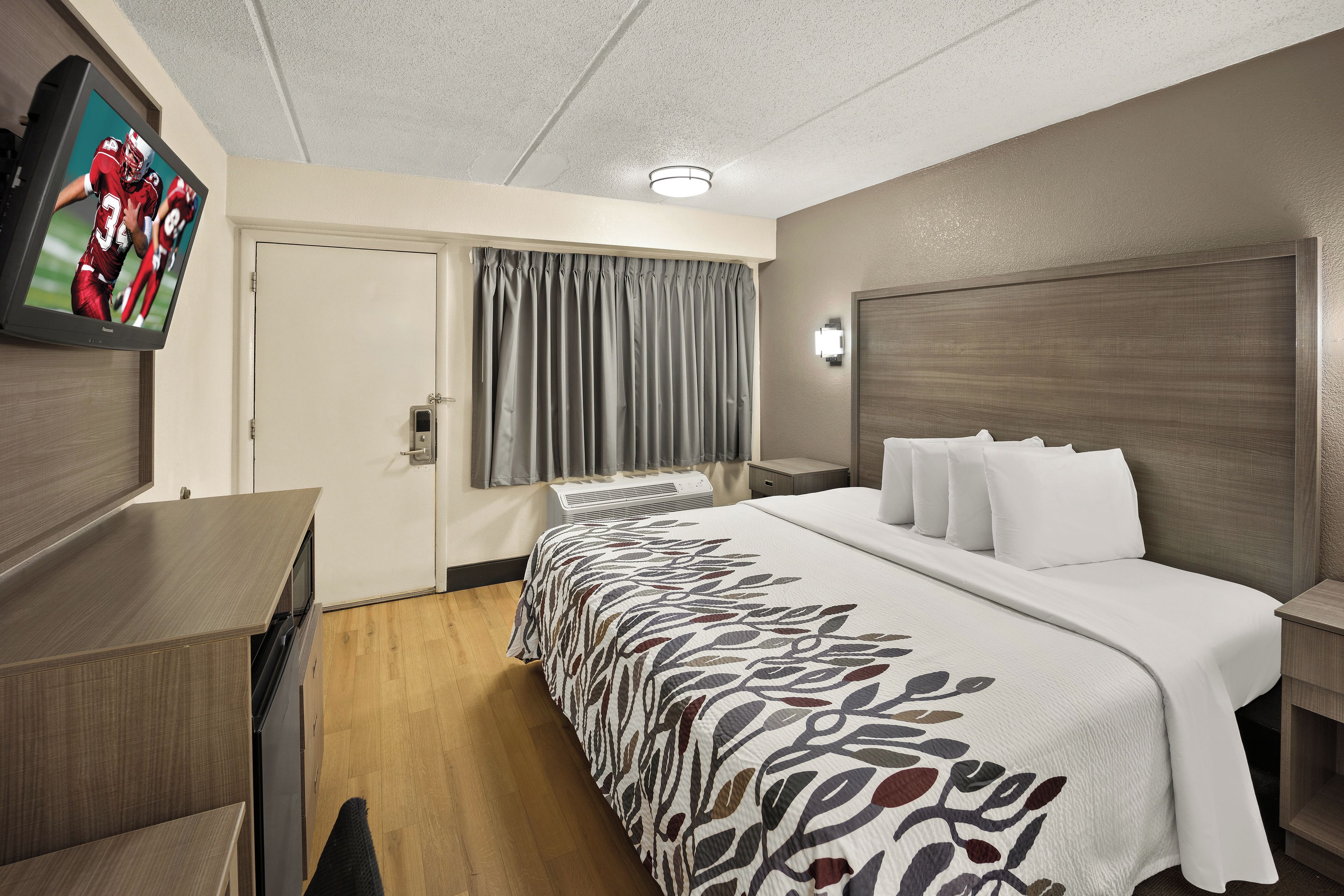 A hotel room with a bed, a TV on the wall, a desk, and a window with curtains.