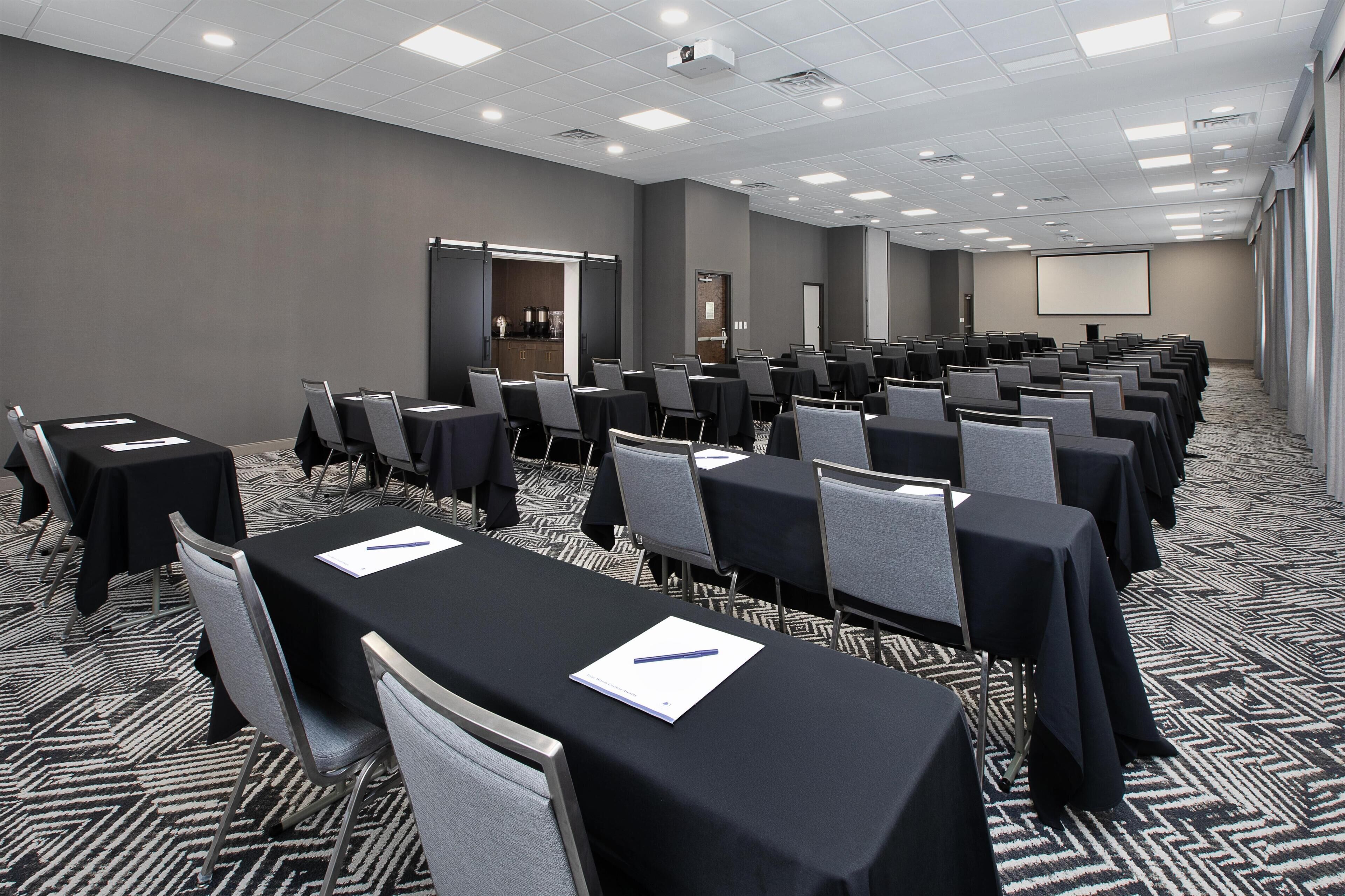 A conference room with rows of tables and chairs, a projector screen, and a door leading to a kitchen area.