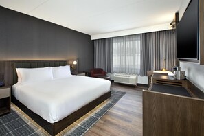 Quarto de hotel com uma cama grande, uma mesa, uma cadeira, uma televisão e uma janela com cortinas.
