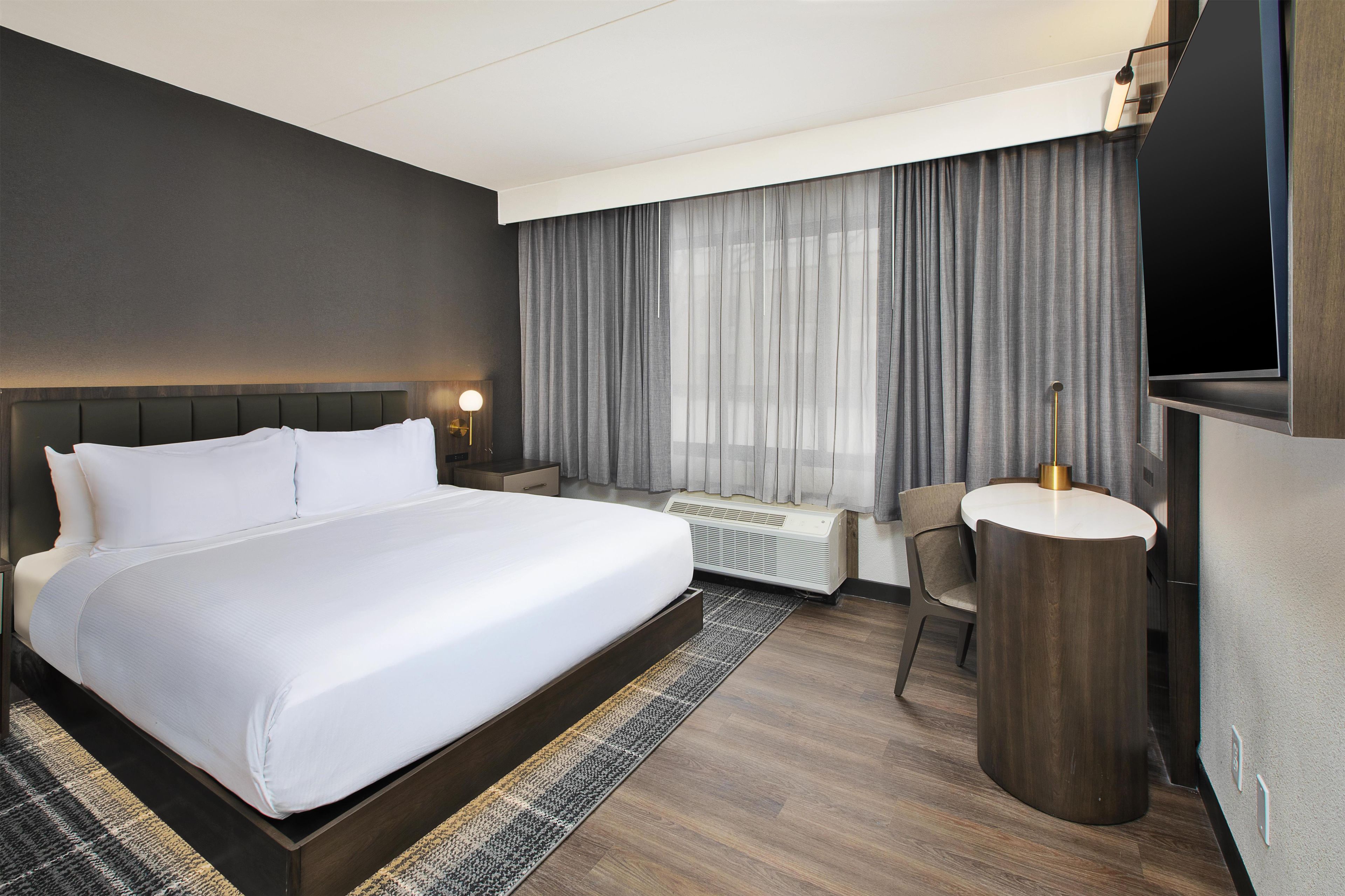 Ein Hotelzimmer mit einem großen Bett, einem Schreibtisch, einem Stuhl, einem Fernseher und einem Fenster mit Vorhängen.