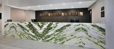 A hotel reception desk with a marble counter, a dark wall with vertical lines, and a modern ceiling with recessed lighting.