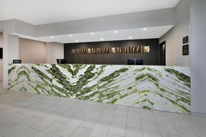 A hotel reception desk with a marble counter, a dark wall with vertical lines, and a modern ceiling with recessed lighting.