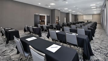 A conference room with rows of tables and chairs, a projector screen, and a kitchenette area.
